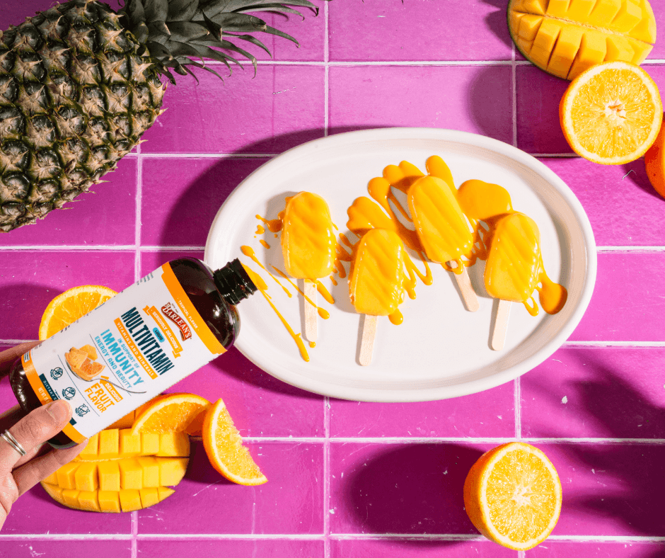 barlean's tropical liquid multivitamin drizzled over mango pineapple popsicles with oranges and mangos and pineapples surrounding them on a purple tiled countertop