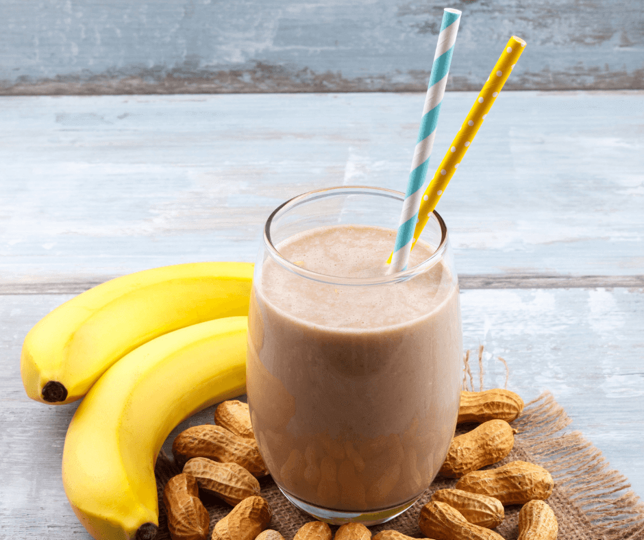 chocolate smoothie with two straws coming out of it sitting on a counter next to bananas and peanuts