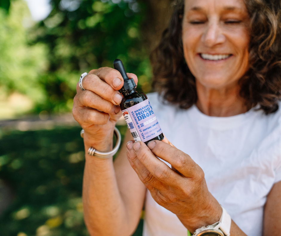 A woman holding a bottle of Mood Drops: Your Secret to Staying Balanced, smiling in a natural outdoor setting.