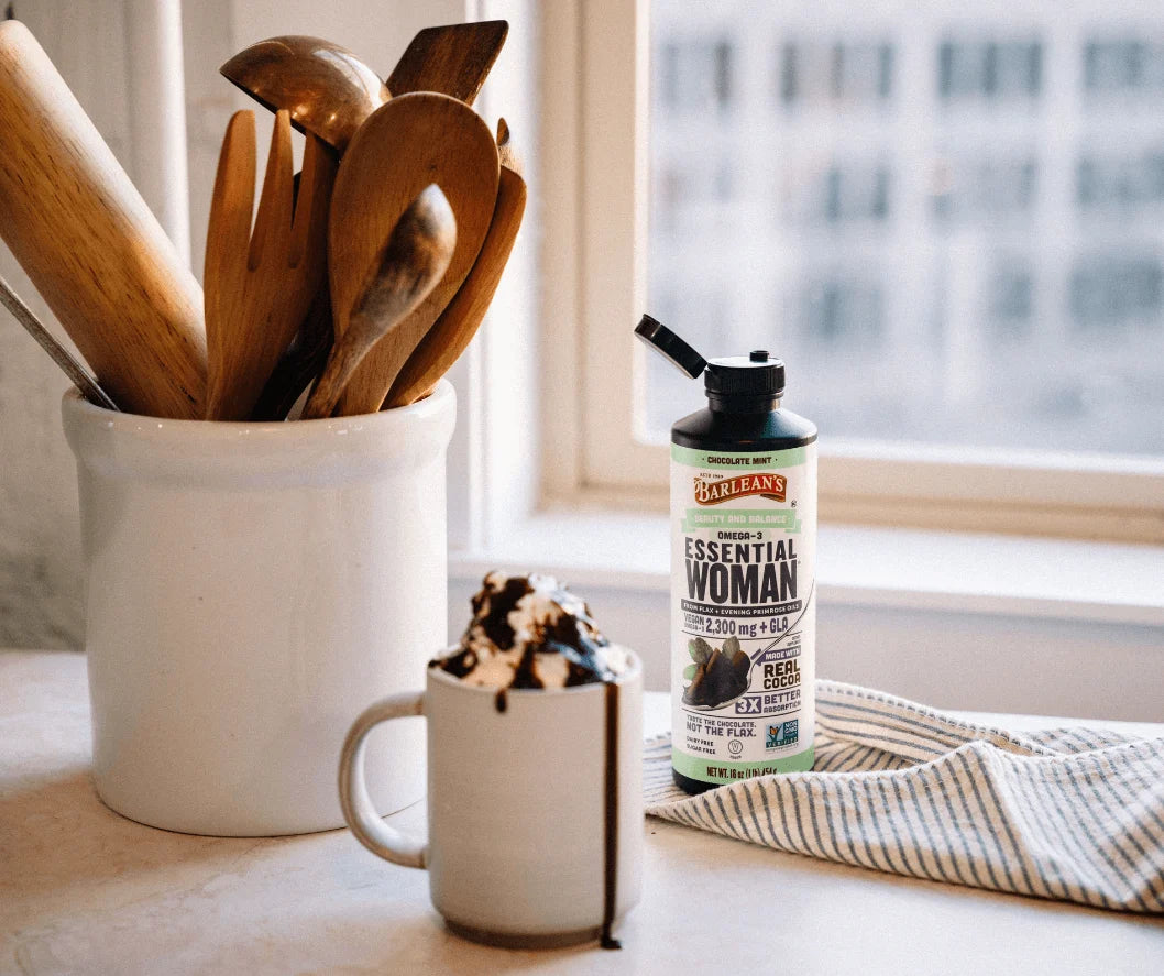 Barlean's Essential Woman supplement bottle next to a coffee cup and wooden kitchen utensils in a bright kitchen.