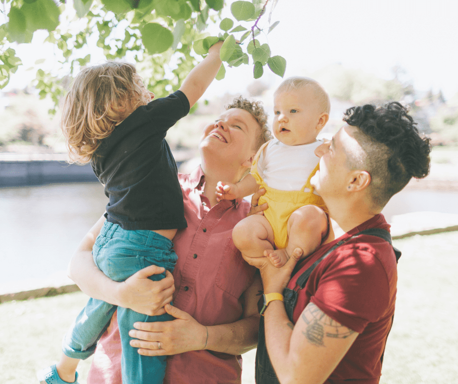 a family of four smiling, the two parents holding their infant and toddler up on their hip and shoulder, the toddler reaching for leaves on a tree
