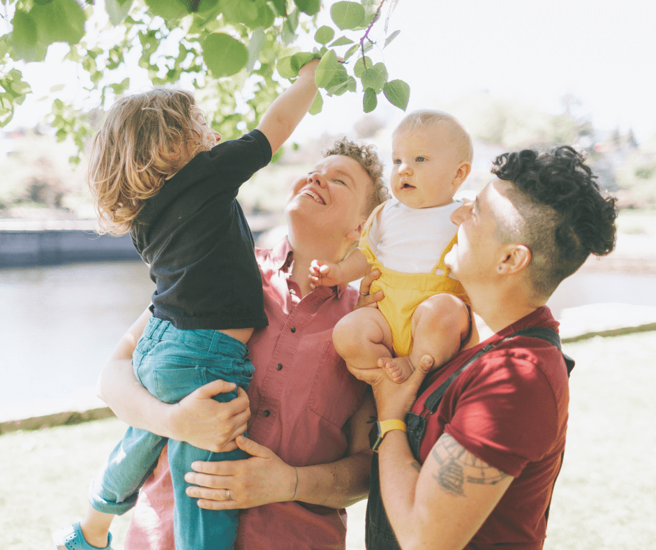 a family of four smiling, the two parents holding their infant and toddler up on their hip and shoulder, the toddler reaching for leaves on a tree