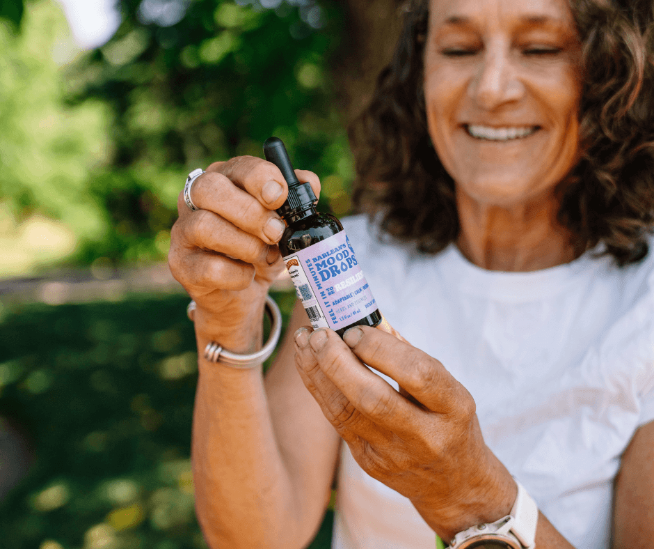 A woman holding a bottle of Mood Drops: Your Secret to Staying Balanced, smiling in a natural outdoor setting.