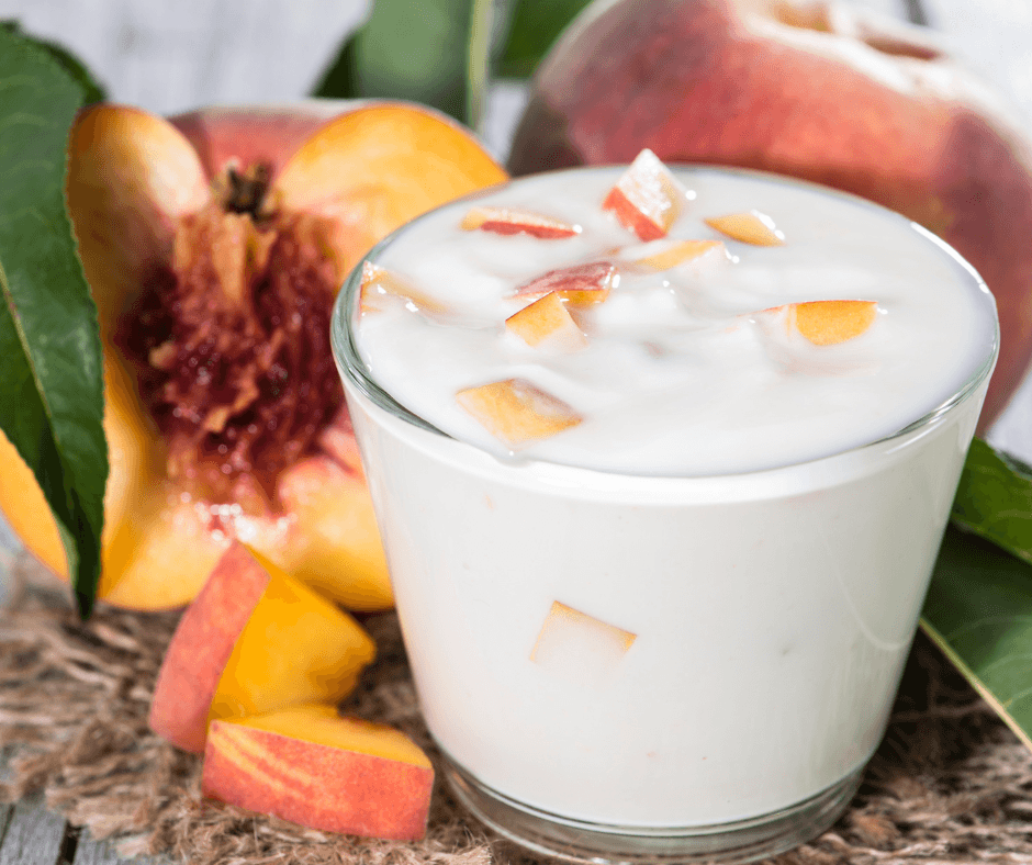 a glass yogurt cup with white yogurt inside and cubes of fresh mangoes and peaches with fresh fruit surrounding