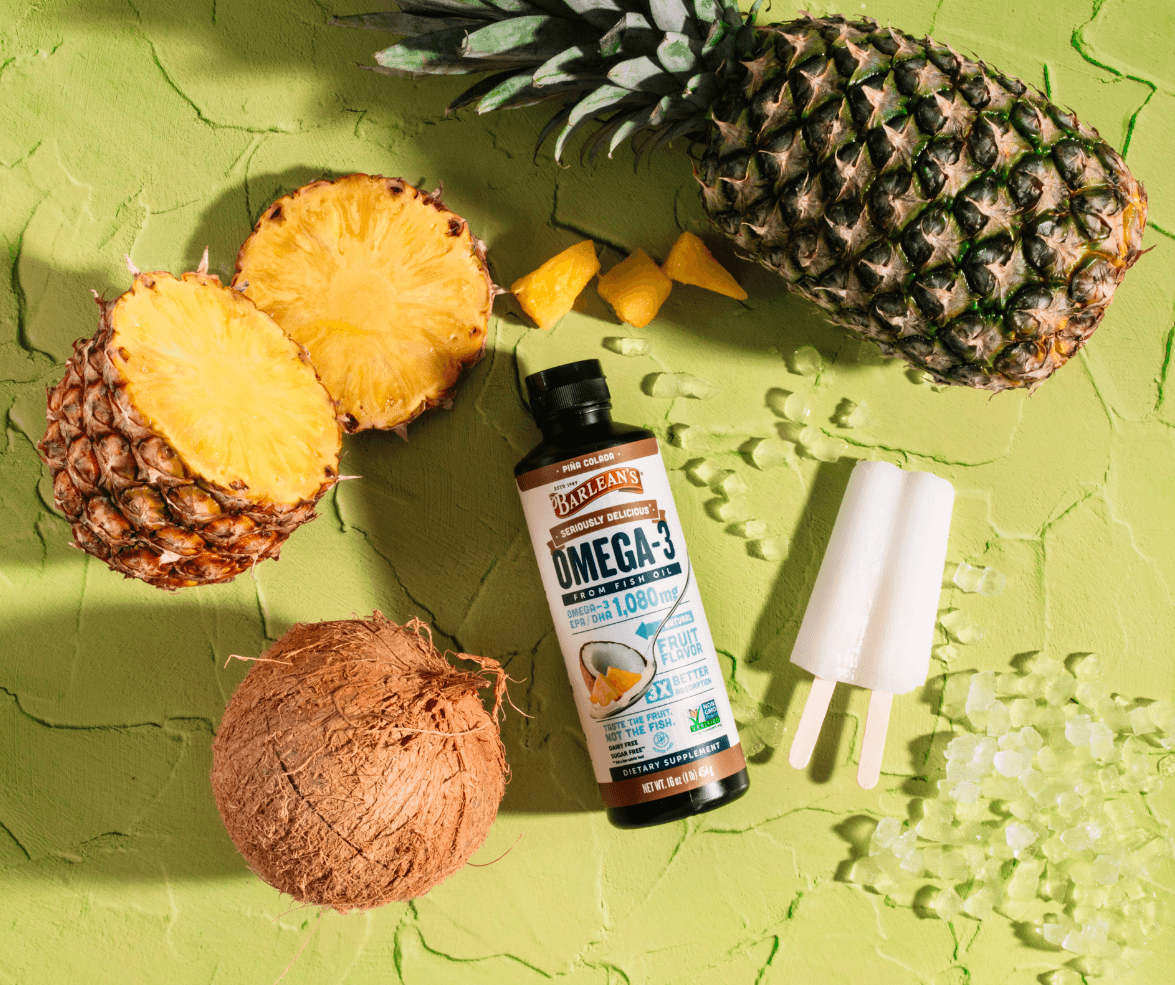 barlean's pina colada smoothie fish oil next to pineapples and coconuts and popsicles and ice cubes on a green countertop