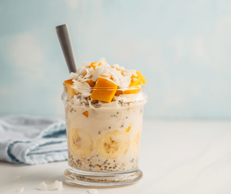 a clear glass with creamy mango peach overnight oats inside with a spoon and topped with fresh mango chunks and coconut shavings